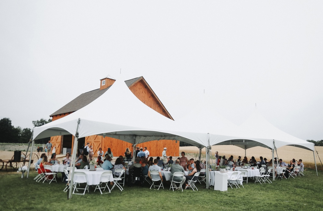 Outdoor wedding with large carnival tent and barn in the background.